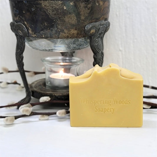 A golden bar of soap stamped with the branding Whispering Woods Soapery on a white surface in front of a vintage warmer with a lit tealight candle.