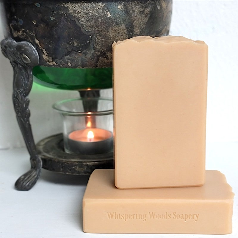 A rectangular bar of soap in a light peach-beige color stands on top of another soap bar. The front bar is stamped with the brand name 'Whispering Woods Soapery.' The soaps stand in front of a vintage warmer with a lit tealight candle.