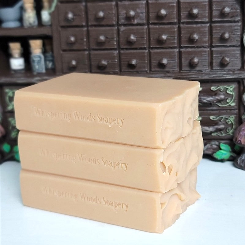 Three stacked rectangular bars of soap in a warm peach-beige color,stamped with the brand name 'Whispering Woods Soapery,' set against a dark apothecary cabinet.