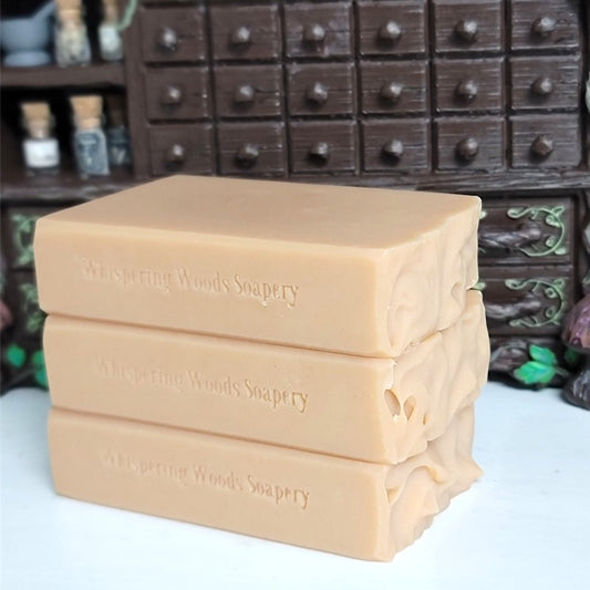 Three stacked rectangular bars of soap in a warm peach-beige color,stamped with the brand name 'Whispering Woods Soapery,' set against a dark apothecary cabinet.