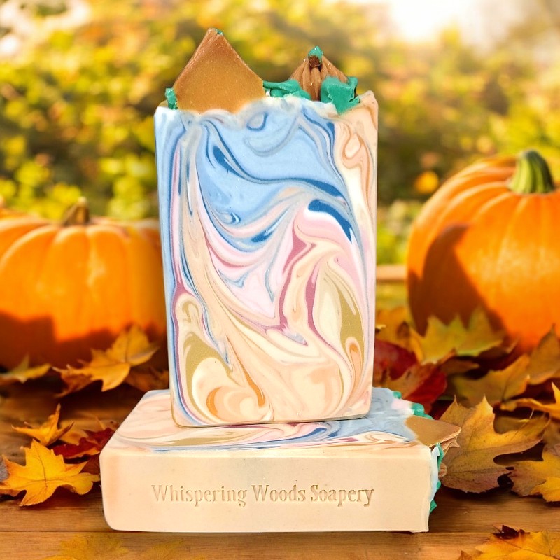 Two soap bars with colored swirls of gold, white, orange, blue and pink, with piped orange pumpkins and green leaves/vines on top, on a wood table with pumpkins and fall-colored leaves