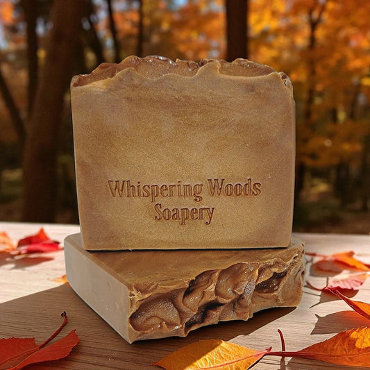 Bars of shimmery brown soap with 'Whispering Woods Soapery' branding on a wooden table with autumn leaves and trees in the background