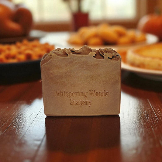 Bar of soap with 'Whispering Woods Soapery' branding on a wooden table with a blurred background of pies and such