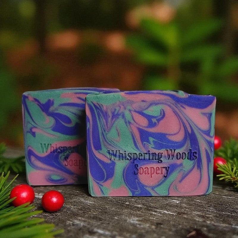 Two colorful soap bars with 'Whispering Woods Soapery' branding on a wooden surface with greenery and berries