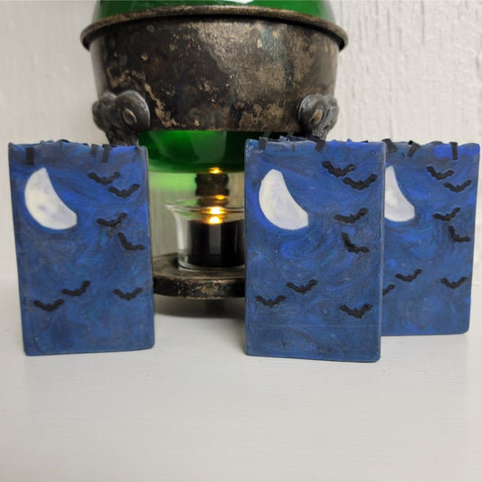 Three rectangular soap bars in shades of dark blue, with small black bats and a crescent moon embed, on a white surface in front of a vintage warmer with a lit tealight candle.