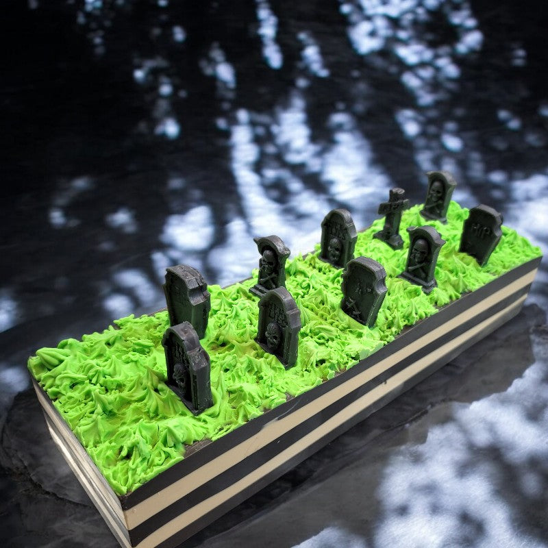 A soap loaf with black and white stripes, topped with neon green grass and tombstones. It sits on a concrete surface with dappled shadows.