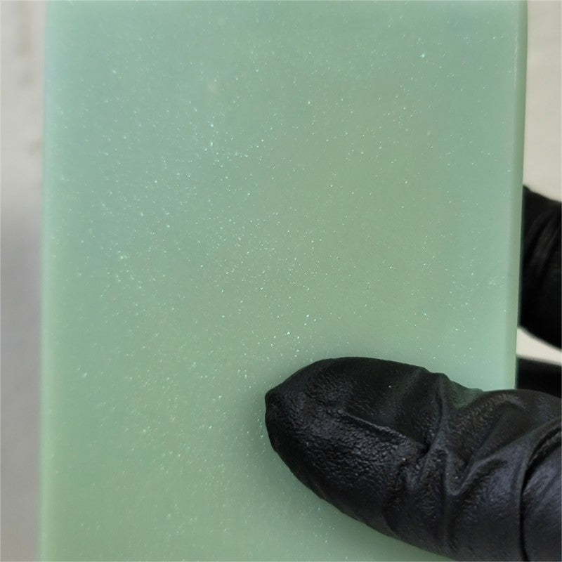 A close-up of a pale green soap bar with many sparkles inside it, held by a black gloved finger at the bottom right.