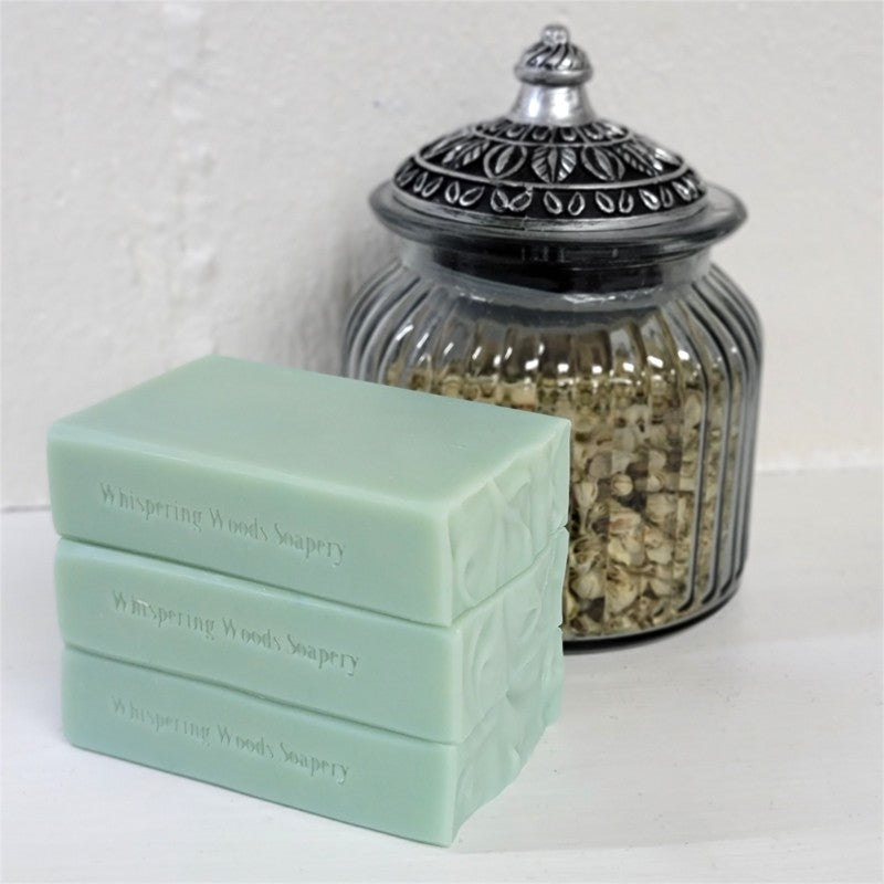 Three stacked bars of pale green soap from Whispering Woods Soapery, displayed next to a decorative ribbed glass jar filled with dried botanicals on a white surface.