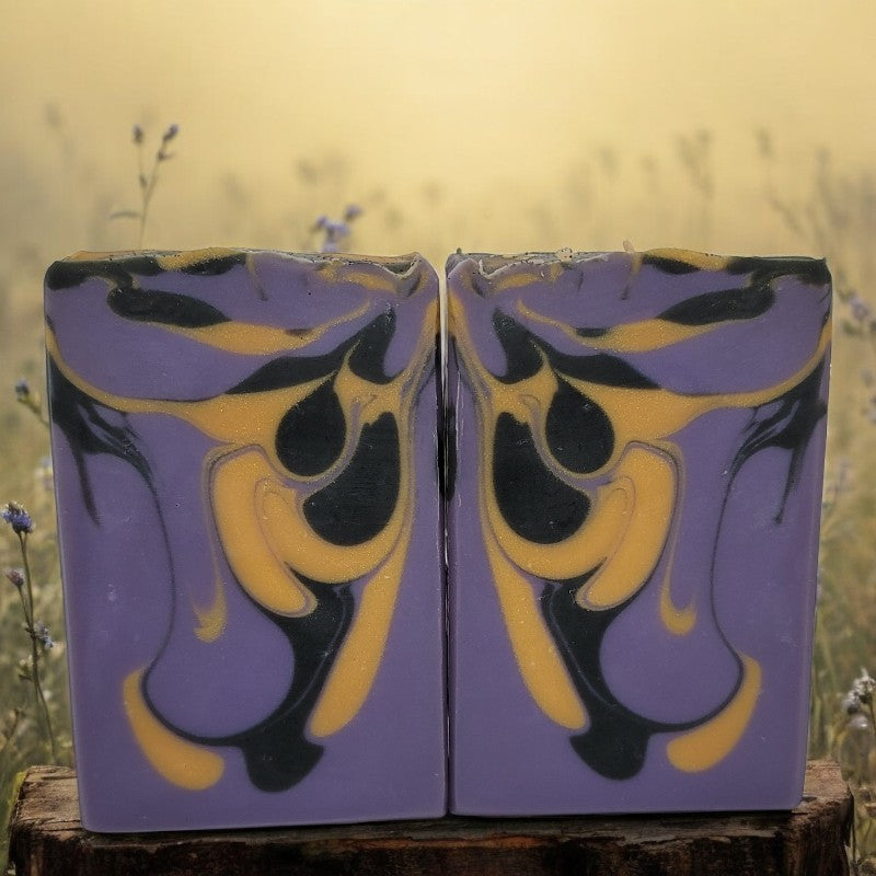 Two soap bars with purple, gold, and black design on a stump with a natural background