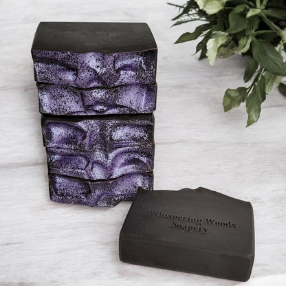 Six bars of black soap with light purple tops are stacked on a marble counter