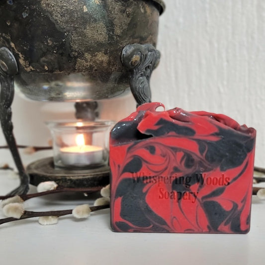 A rectangular bar of soap with a red and black marbled swirl pattern sits in front of a vintage metal warmer and a small burning candle. The soap is stamped with the text 'Whispering Woods Soapery'.