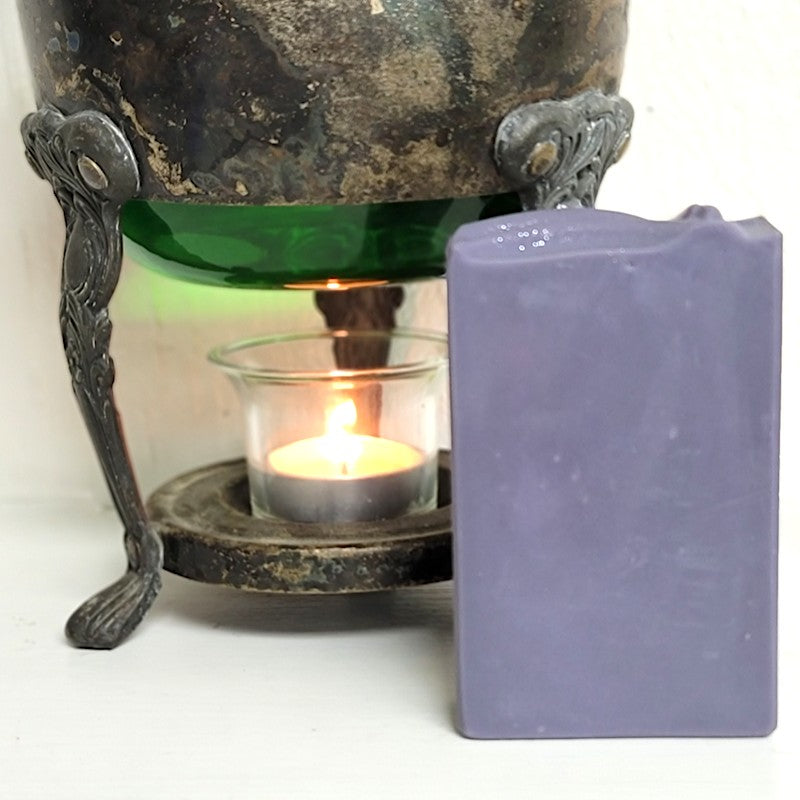 A purple bar of rectangular soap in front of a vintage warmer with a lit tealight candle on a white surface.