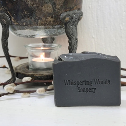 A rectangular bar of light black soap from Whispering Woods Soapery, stamped with the brand name, styled in front of a vintage warmer with a lit tealight candle.