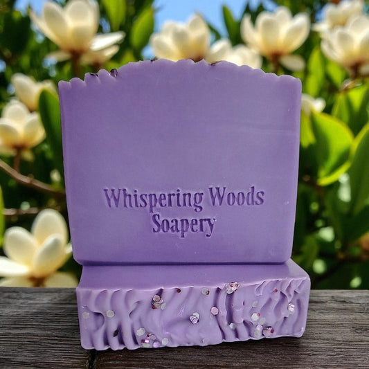 Purple soap bars with 'Whispering Woods Soapery' text against a natural background of magnolias
