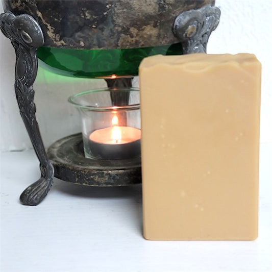 A rectangular bar of pale orange soap stands next to a small lit tealight candle in a dark metal vessel with a green interior on a white surface.