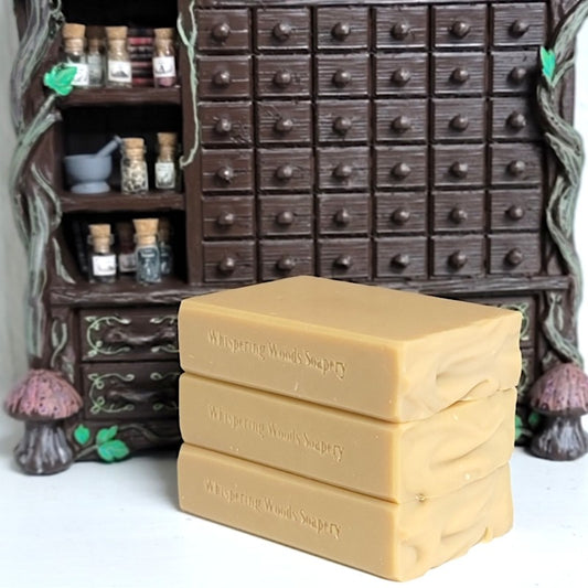 Three stacked bars of pale orange soap labeled 'Whispering Woods Soapery' in front of a decorative dark brown miniature apothecary-style cabinet with small bottles.