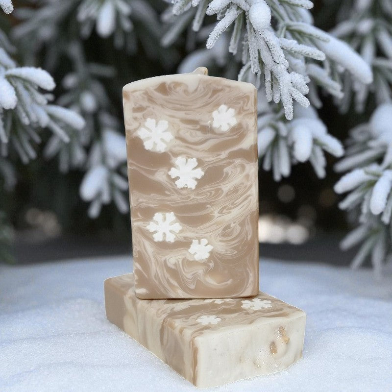 Two cream and toffee colored soap bars with little snowflake embeds on a snowy surface with snow-covered trees in the background