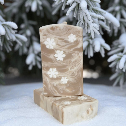 Two cream and toffee colored soap bars with little snowflake embeds on a snowy surface with snow-covered trees in the background