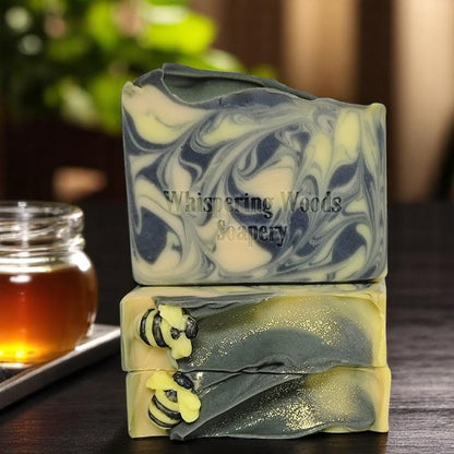 Three bars of soap with yellow, gold and black swirls that have tiny soap bees on top sit on a dark table with a jar of honey