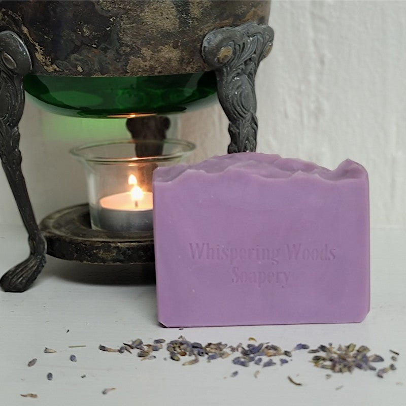 A lavender-colored bar of soap from Whispering Woods Soapsy stands on a white surface in front of a rustic metal candle holder with a lit tealight candle. Scattered dried lavender buds are visible in the foreground.