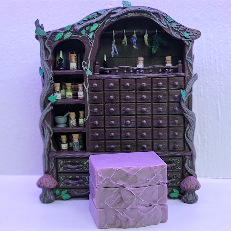 A miniature fantasy-style wooden alchemist's cabinet with shelves of miniature potion bottles, hanging herbs, and a large front drawer compartment, shown with a stack of lavender soap in front on a white surface against a pale purple background.