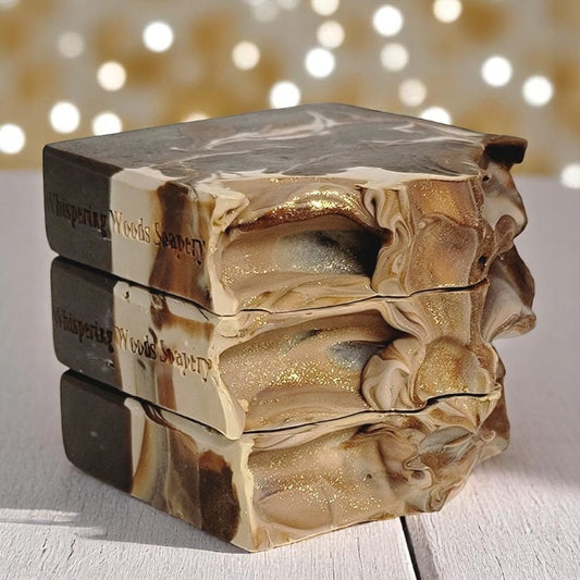 Three bars of soap with cream, caramel and brown swirls are stacked on a wood surface with soft gold lighting in the background