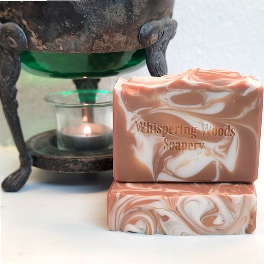 Two rectangular artisan caramel-colored soap bars with a white marble swirl pattern are stacked on a white surface. The top bar is stamped with the brand name 'Whispering Woods Soapery.'