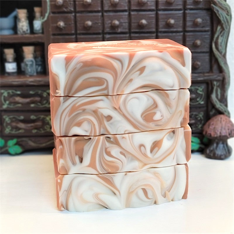 Four stacked rectangular bars of handmade soap with a marbled swirl pattern in caramel and white, set against a blurred background of a brown apothecary cabinet.