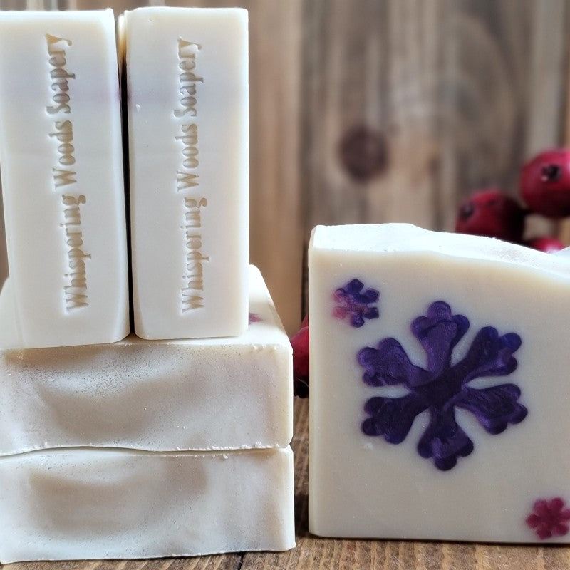 Five bars of white soap, one showing a large purple snowflake in the center, sit on a wood surface with berries in the background