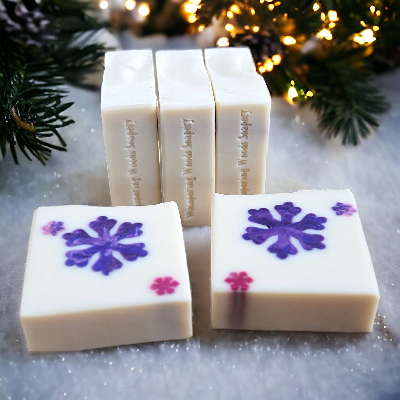 Five bars of white soap with a big purple and pink snowflake in the middle lay on the snow with lighted pine branches in the background