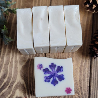 Five bars of white soap, one with a large purple snowflake in the middle, lay on a wood surface with pinecones