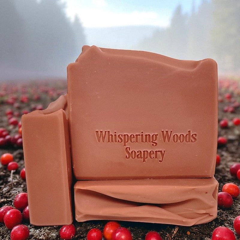 Bars of dusty pink soap with 'Whispering Woods Soapery' branding in a natural setting with red berries