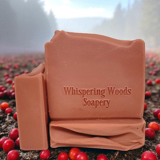 Bars of dusty pink soap with 'Whispering Woods Soapery' branding in a natural setting with red berries