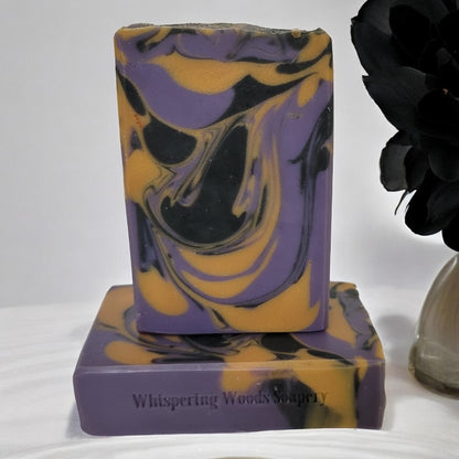 Two bars of purple, gold and black swirled soaps on a white cloth with black feathers in a vase beside them