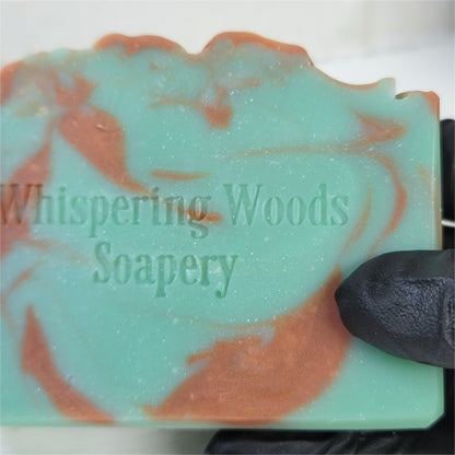 A close-up of a handmade soap bar with a bluish-green sparkly base and copper swirls, held by a black gloved hand. The soap is stamped with the brand name 'Whispering Woods Soapery.'