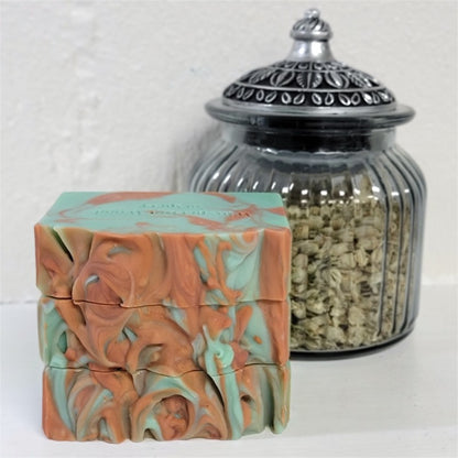 A stack of three bluish-green soap bars with copper swirls displayed next to a glass jar filled with dried botanicals on a white surface.