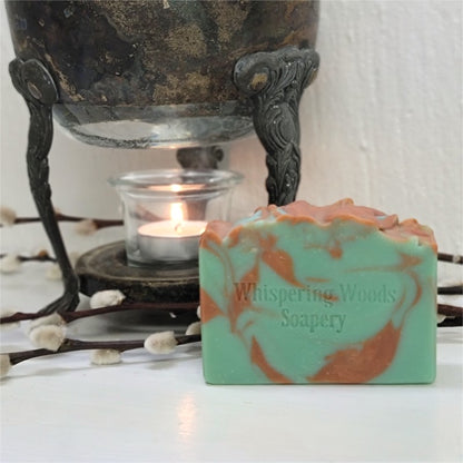 A bluish-green rectangular bar of soap with copper swirls sits in front of a vintage warmer with a lit tealight candle. The soap is stamped with the brand name 'Whispering Woods Soapery'.
