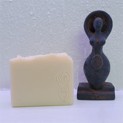 A cream-beige bar of soap with a stamp of the divine goddess in the right corner sits by a brown wax figure of the spiral goddess on a white background.
