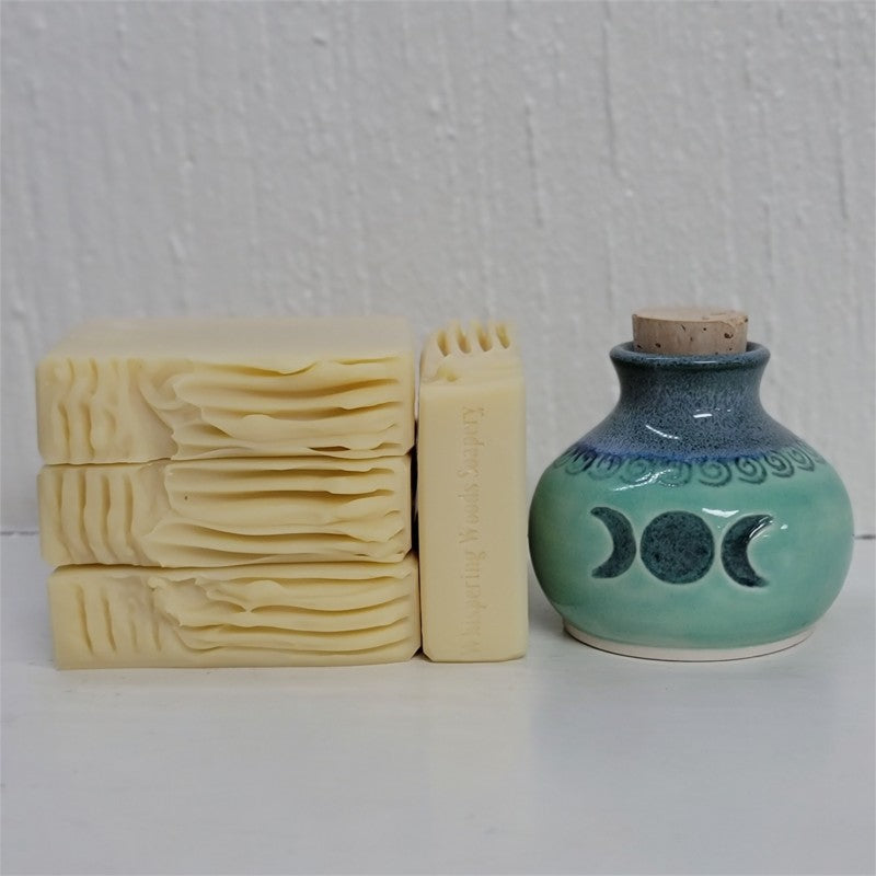 Four cream-beige bars of soap, one with the stamp of 'Whispering Woods Soapery,' sit by an artisan crafted clay jar in shades of teal and blue, with the triple moon symbol on its front. The background is white.