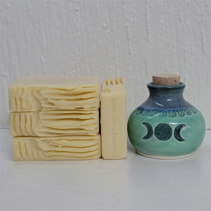Four cream-beige bars of soap, one with the stamp of 'Whispering Woods Soapery,' sit by an artisan crafted clay jar in shades of teal and blue, with the triple moon symbol on its front. The background is white.