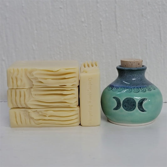 Four cream-beige bars of soap, one with the stamp of 'Whispering Woods Soapery,' sit by an artisan crafted clay jar in shades of teal and blue, with the triple moon symbol on its front. The background is white.