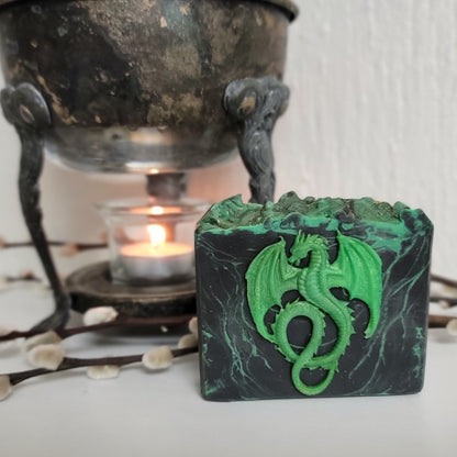 A bar of dark green and black marbled soap, with a raised green dragon design on the front, next to a burning candle in a vintage warmer.