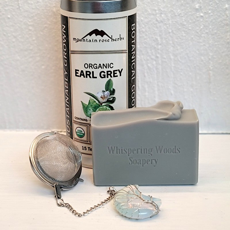 A gray bar of soap, stamped 'Whispering Woods Soapery, on a white surface next to a tin of Earl Grey tea and a silver tea strainer with a wire-wrapped crystal moon.