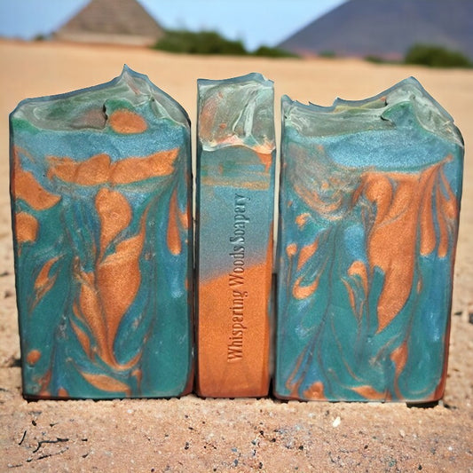 Three turquoise and coppery-colored bars of soap sitting in sand with pyramids in the background