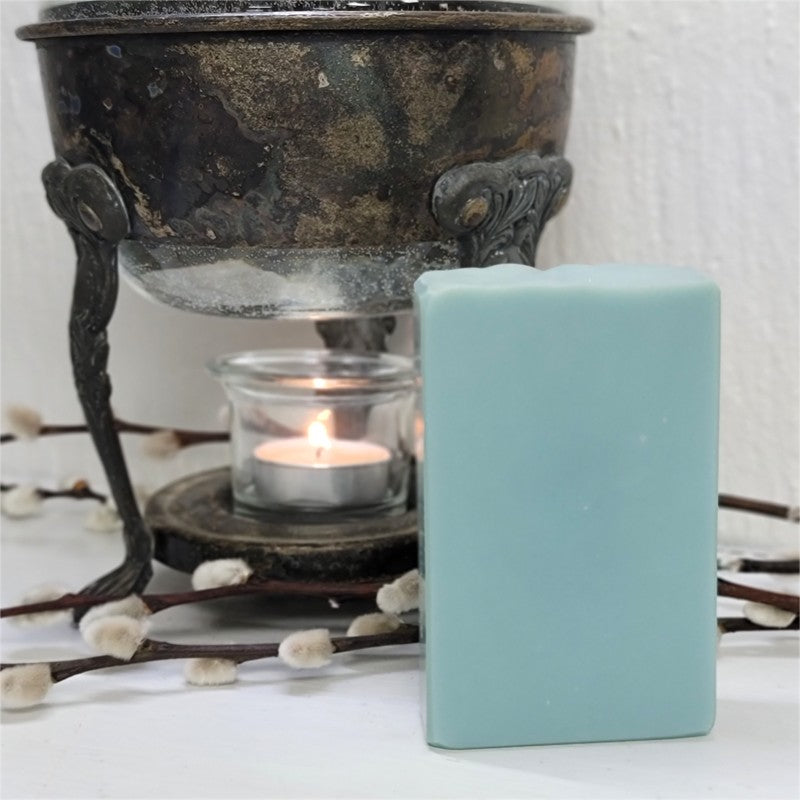 A duck-egg-blue bar of soap from Whispering Woods Soapery stands on a white surface with a vintage warmer with a lit tealight candle in the background.