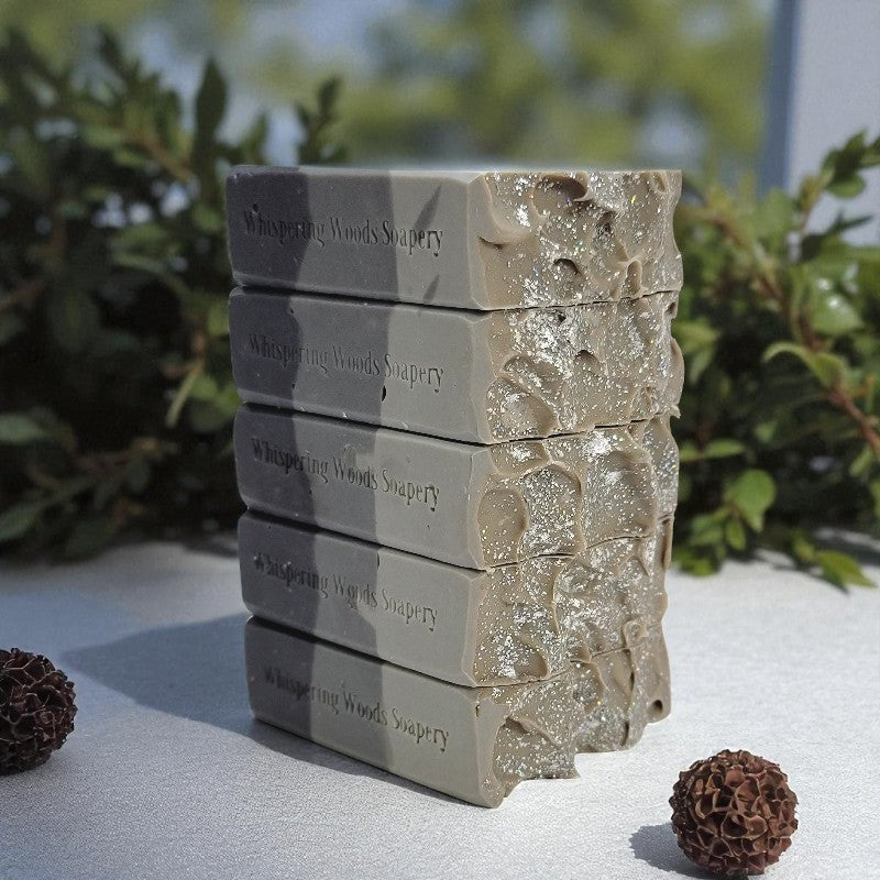 Five bars of soap labeled 'Whispering Woods Soapery' stacked on a white surface with garland behind them