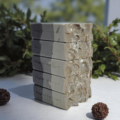 Five bars of soap labeled 'Whispering Woods Soapery' stacked on a white surface with garland behind them