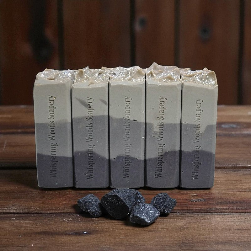 Five bars of soap with a gradient from gray to black, branded 'Whispering Woods Soapery', on a wooden surface with lumps of coal