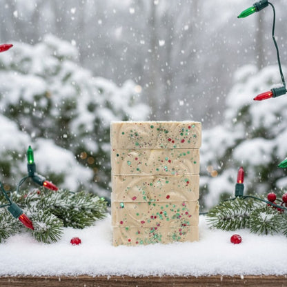 Five bars of soap with green and red sparkles stacked on a wood surface with falling snow and green and red christmas lights
