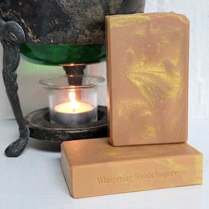 Two rectangular bars of orange soap with gold sparkly swirls, one standing upright and one lying flat with 'Whispering Woods Soapery' stamped on it. A lit tealight candle in an antique glass warmer is in the background.